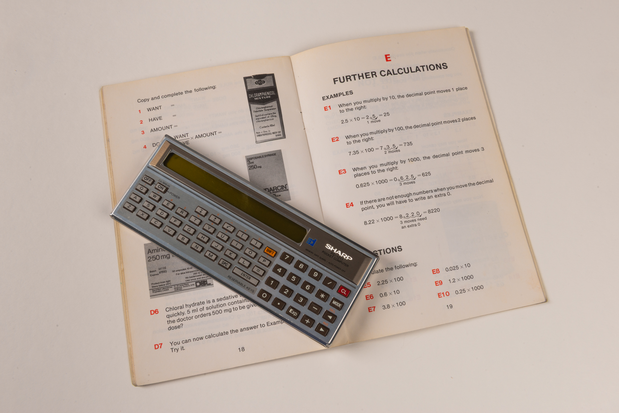 An open mathematics workbook underneath a flat, rectangular calculator with a silver body and a golden-coloured digital display.