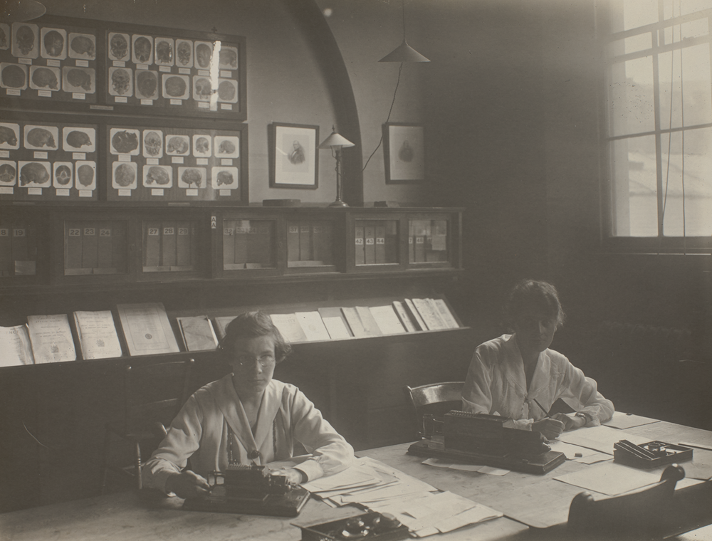 Greyscale image of two women working at a table in the Galton Laboratory. They have papers and equipment in front of them and there are shelves and books lining the wall behind them.