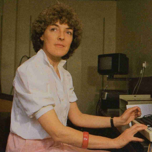 Sue Norman, a white woman with short, light brown, curly hair, a white shirt, and pink trousers sits at a desk with her hands on a computer keyboard. In the background a boxy TV/computer monitor and cables are visible.