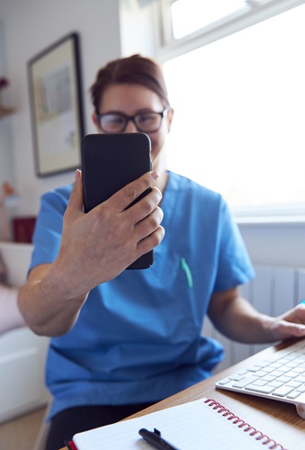 A white, woman nurse with brown hair and glasses and wearing blue scrubs. She is sitting at a desk with a computer keyboard and notepad visible, she is holding up a smartphone in front of her face as though she is on a video call with a patient.