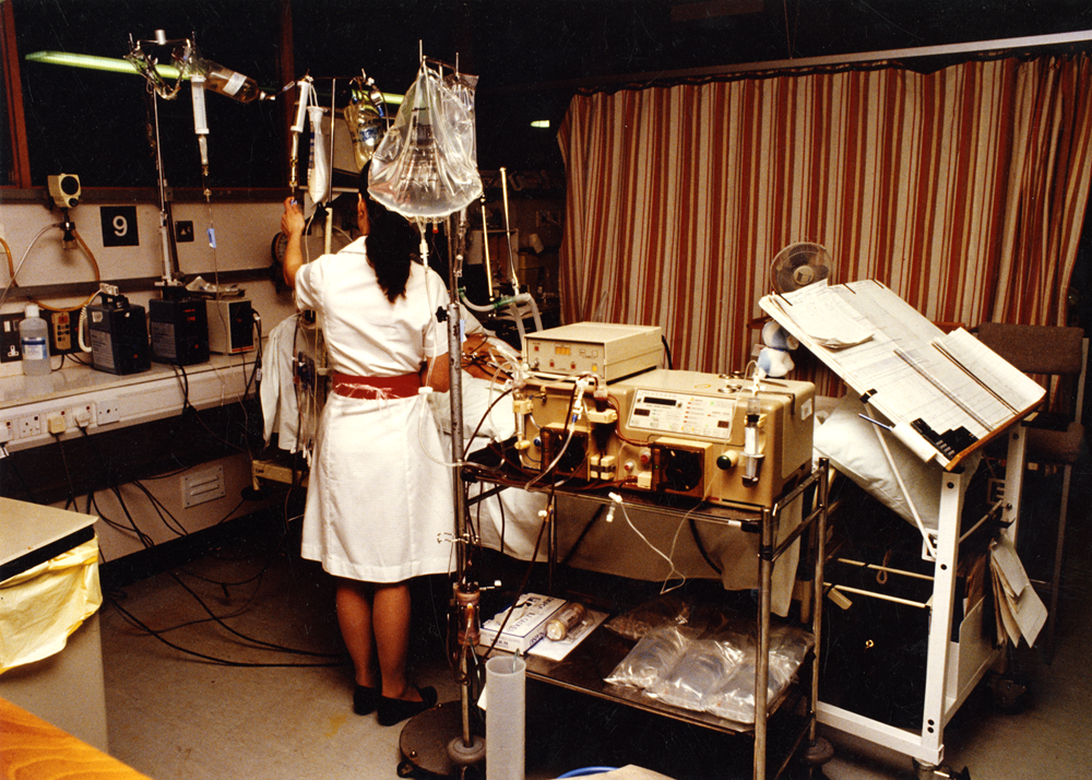 A 1990s hospital room with a woman nurse in a white dress and red belt checking equipment attached to a patient. There are medication drips, bulky electric monitoring equipment, paper monitoring charts and in the background is a brown and beige striped curtain to separate the next bed.