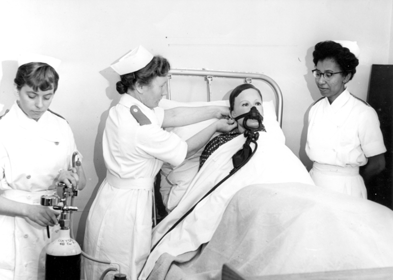 Greyscale image of three 1950s student nurses training using a dummy. One nurse is handling an oxygen tank, the second is attaching a breathing mask to the dummy and the third is standing on the other side of the bed observing.