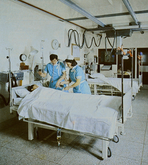 Two 1980s nurses in blue dresses and white caps tending to the equipment around a patient who is lying in bed. There are many tubes and wires in the background, and various equipment such as oxygen and a catheter is visible.