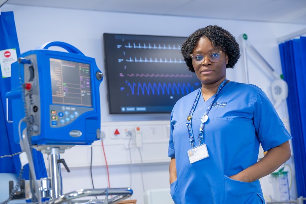A black, woman nurse wearing glasses, blue scrubs and a lanyard stands in a hospital - next to and behind her are monitoring machines.