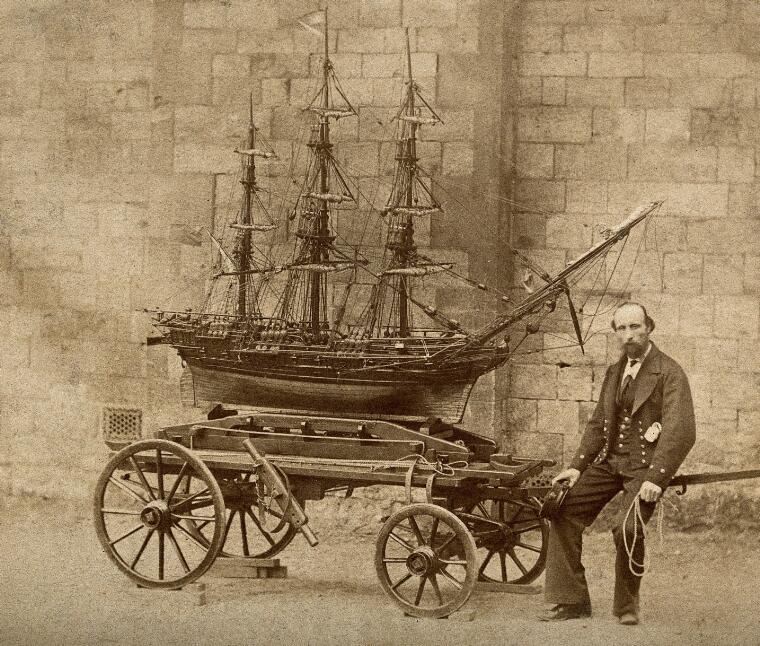 Sepia photograph of James Pullen, sitting on the end of a wooden trolley upon which rests a huge model ship made by him.