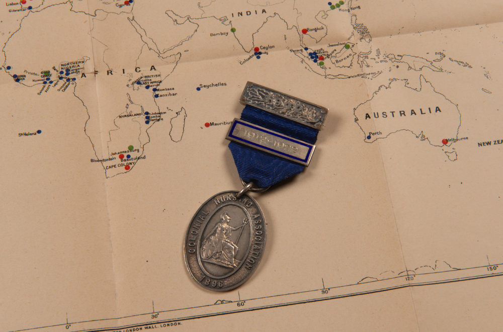Colonial Nursing Association badge with the dates 1912-1922 etched in one bar, silver metal pieces connected with a royal blue coloured ribbon. The main ovular badge shows a relief image of a seated Britannia holding a trident, shield, and wearing a plumed helmet. The badge is laid on top of a map, the close-up section of which shows Africa, Australia and some of Asia with particular locations marked with a green, red or blue dot.