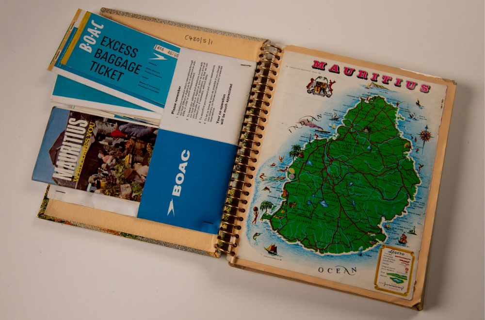 A scrapbook laid open on a white-grey background. The double page spread contains a BOAC (British Overseas Airways Corporation) pouch which contains a 'Mauritius welcomes you' pamphlet and a BOAC excess baggage ticket, as well as other papers that are minimally visible underneath. On the right side page a colourful, glossy map of Mauritius has been glued in.