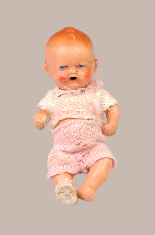 An historic baby doll with pink-peach coloured skin, blue eyes and blonde hair, dressed in a frayed pink and white knit outfit with one bootie missing.
