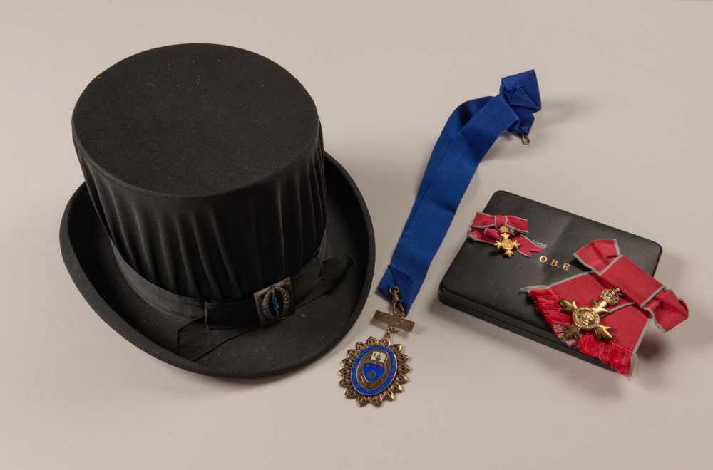 A black top hat with a black ribbon, and metal badge above rim resting next to a blue ribbon with an RCN 'Fellow' medallion and a black box on which rests two red and gold OBE awards.