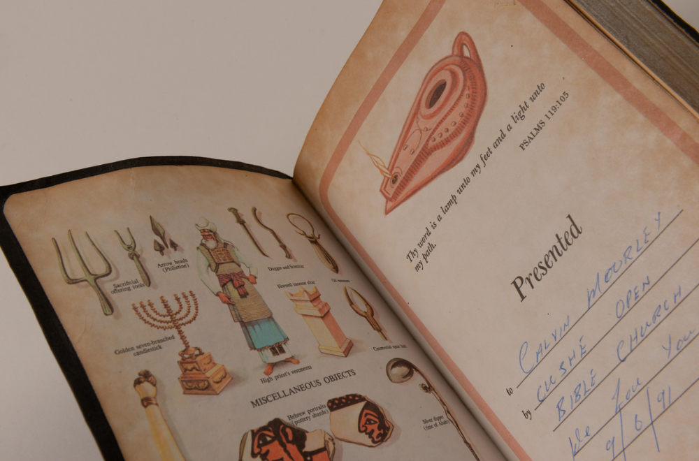 Front page of an opened Bible displaying colour illustrations of ancient religious items including sacrificial offering tools, dagger and scimitar, arrow heads, candlestick, vestments, and altar on the left page. On the right page is an illustration of an oil lamp with the quote 'Thy word is a lamp unto my feet and a light unto my path' (Psalms 119:105) and a message that the Bible was presented to 'Calvin Moorley by CUSHE Open Bible Church' on the 9th June 1991.