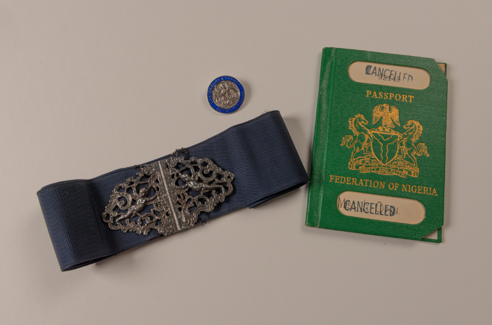 A blue nurse's belt with an ornate silver buckle, a bright, green Nigerian passport stamped "CANCELLED," and a circular silver and blue, enamelled badge for Canterbury & Thanet School of Nursing are displayed on a light background.