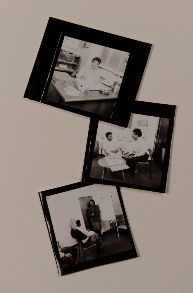 Three black-and-white photographs showing mostly black women working in the Sickle Cell Unit at St Bartholomew's Hospital in 1987. In the top image one woman sits at a desk holding a pen, smiling at the camera and surrounded by papers and other office equipment. The second image shows two women sitting in chairs behind a small round table with a folder laying open; they are facing each other and smiling. The third image has a black woman standing next to a flipchart and talking to a group of seated women facing away from the camera.