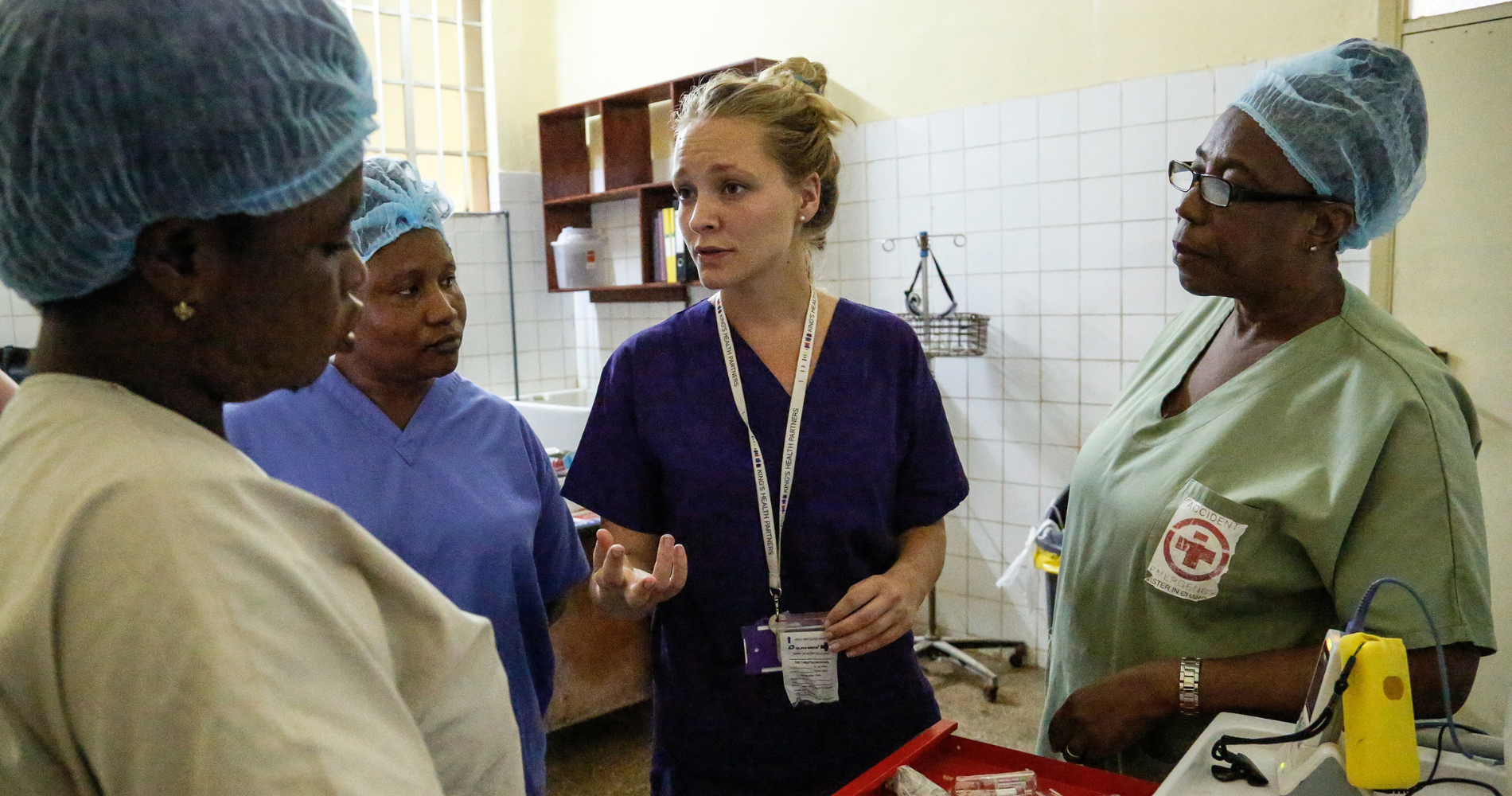 A group of four nurses stand in a hospital room in Sierra Leone. They wear scrubs and three have surgical caps, engaged in a discussion. Medical equipment is visible in the foreground and background.