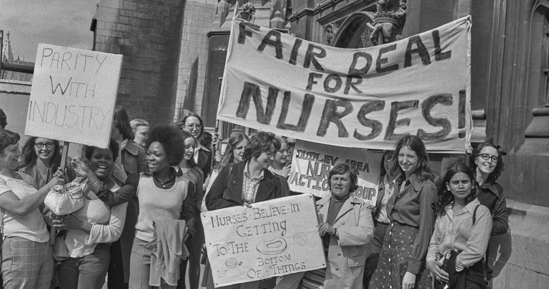 A greyscale image of a group of nurses holding banners reading "Fair Deal for Nurses", "Parity with Industry", and "Nurses believe in getting to the bottom of things" in a protest for fair pay.