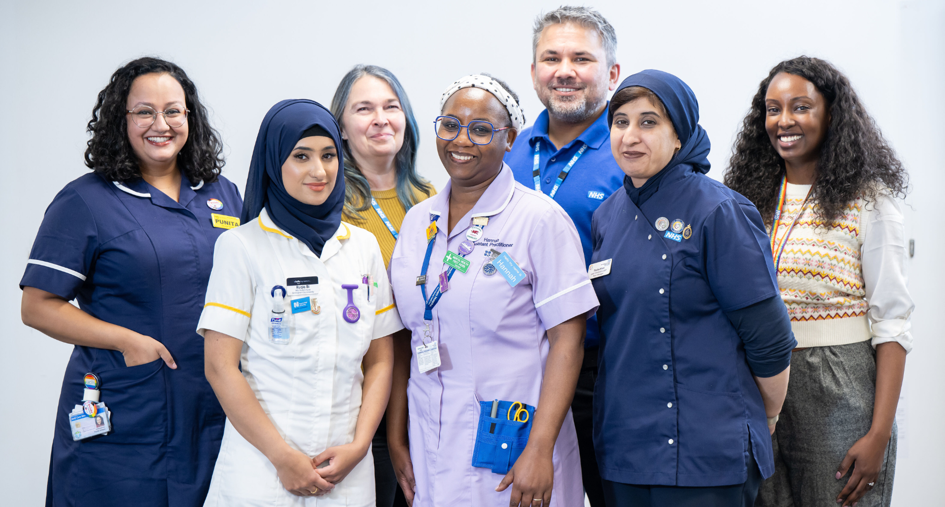 An ethnically diverse group of seven healthcare professionals, including both men and women, standing together and smiling at the camera in a brightly lit indoor setting.