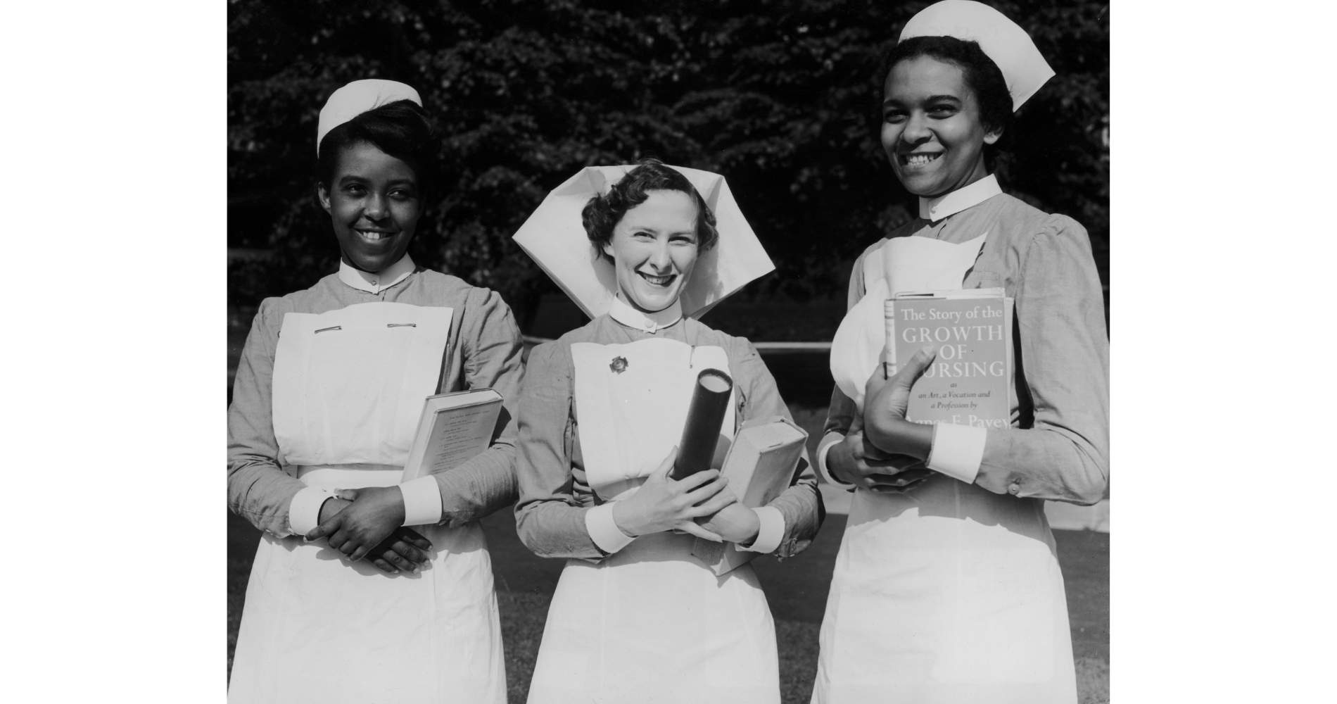 Three 1950s nurses, all of different ethnicities, in uniforms including caps, dresses and aprons stand next to each other smiling at the camera, holding books, in a garden setting.