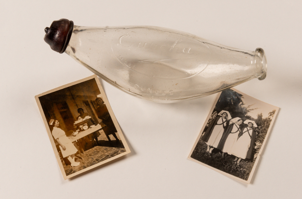A clear, glass banana-shaped baby bottle with a dark rubber teat at one end. Placed just in front of the bottle are two small images - the left one shows three nurses and a baby being weighed on scales, and the right one shows three nurses in full uniform including dark, woollen capes, who are looking at the camera. Except for the middle nurse, Rosa Cassels, who is looking to the left so only the right side of her face is visible.