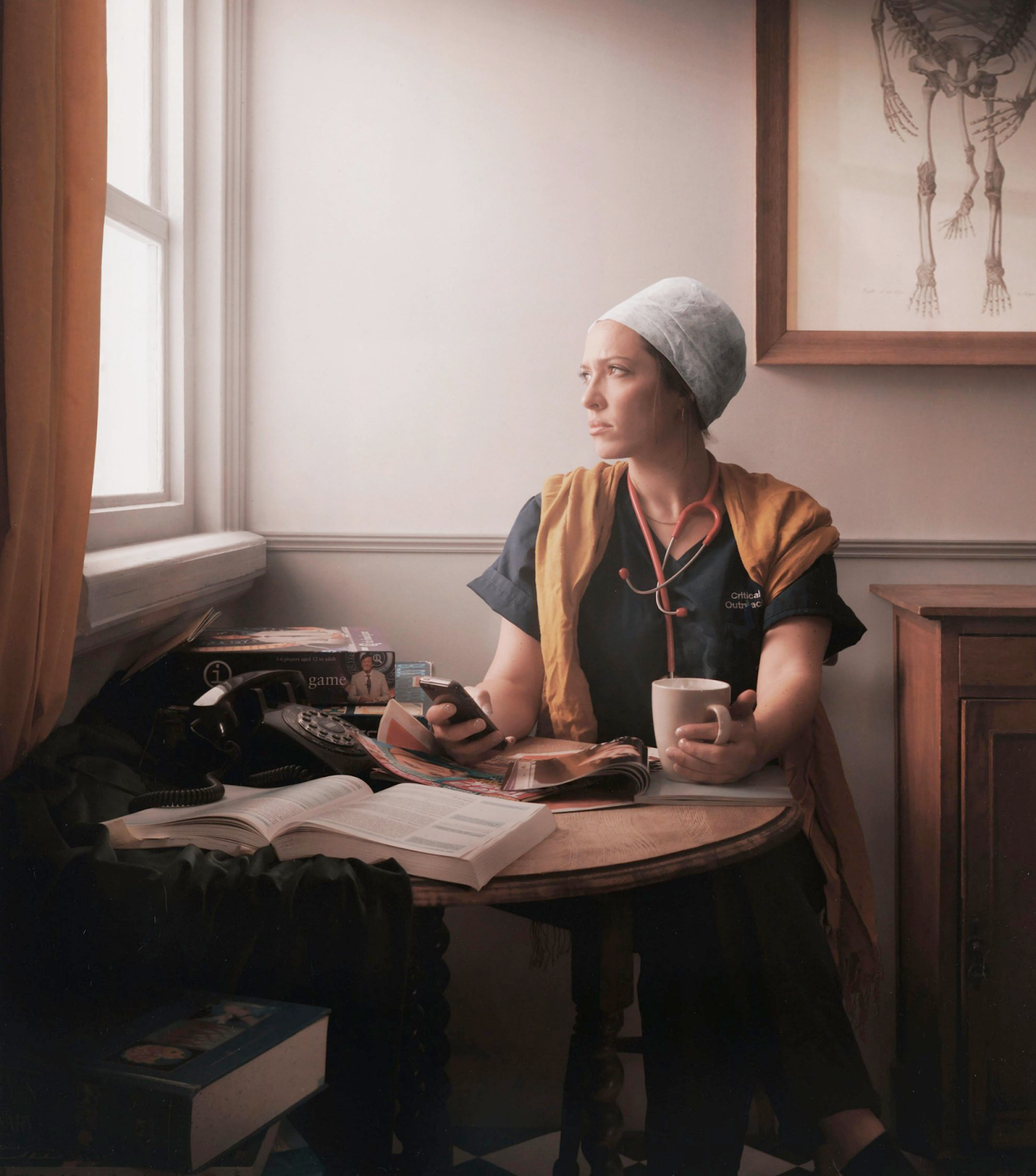 Photograph of a nurse sitting at a round table covered with books, magazines and a dial phone, holding a cup of coffee in one hand and a phone in the other, looking out of the window.