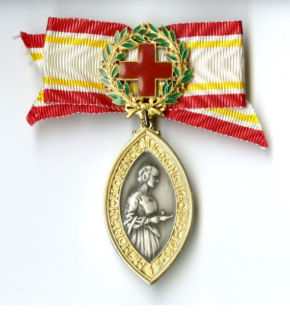 An oval gold and silver badge with a central silver image of Florence Nightingale surrounded by a gold banner containing possibly Latin writing. This is suspended from a green wreath surrounding a red cross. This is all attached to a gold, white and red ribbon