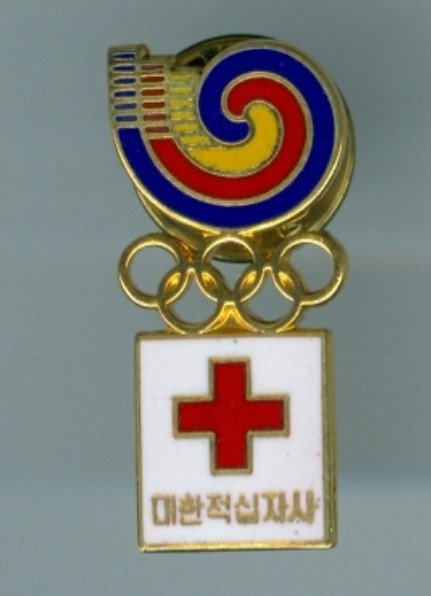 A colourful gold and enamel badge, with a blue, red and yellow spiral at the top. Underneath are the five Olympic rings and hanging from this is a white square with a red cross