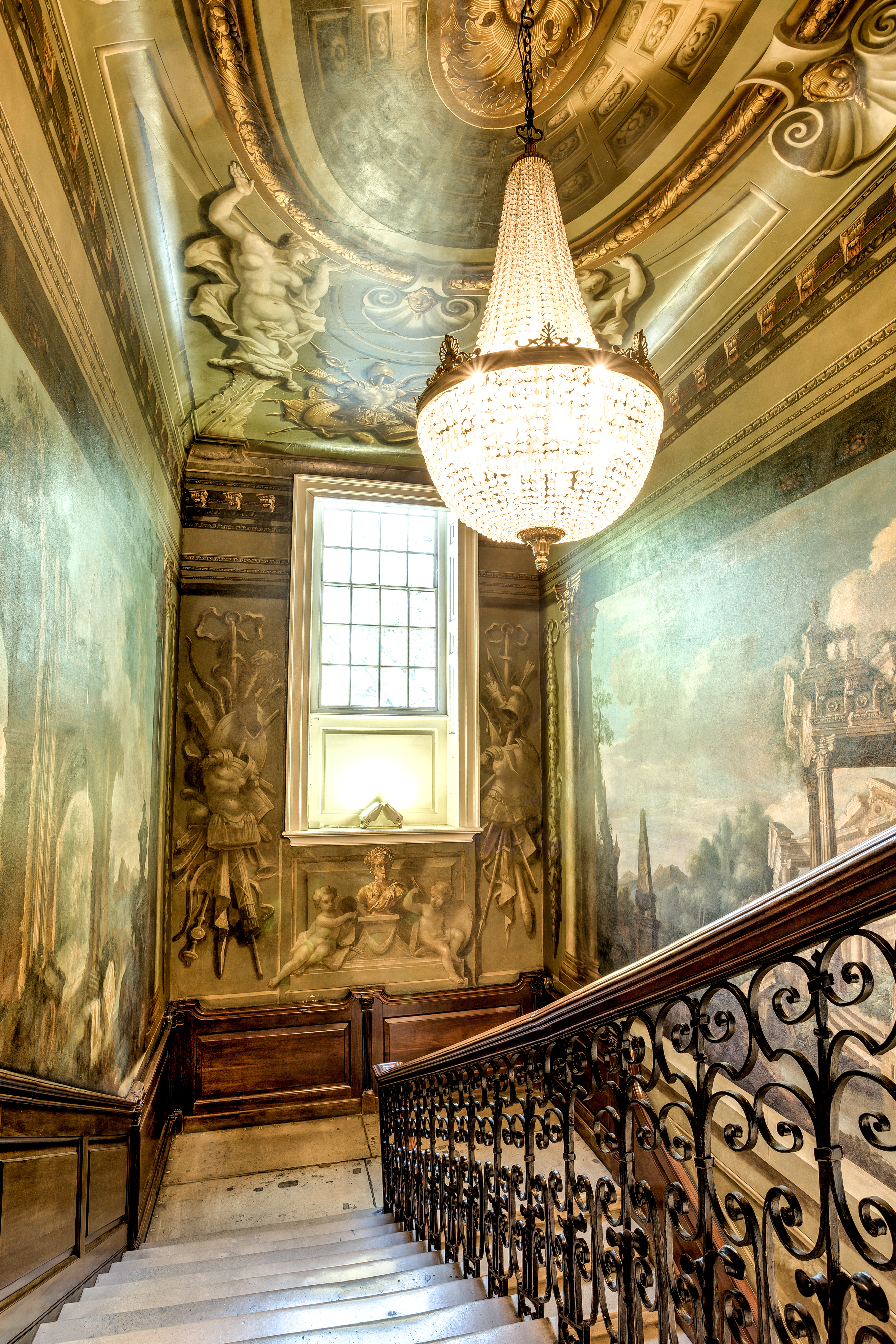 Highly decorative staircase viewed from the top with a lit, pear-shaped chandelier, openwork metal railings supporting a wooden banister. The walls are painted in a baroque style depicting Greek architecture and figures.