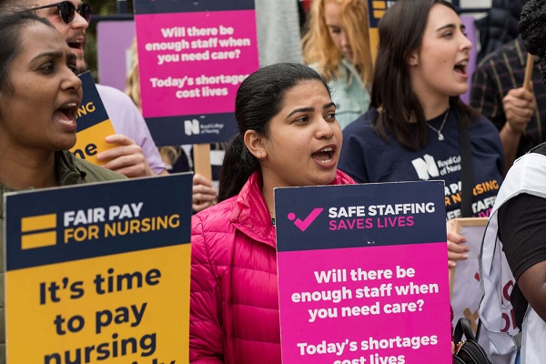 Nursing staff at a demonstration holding signs saying 'Fair Pay For Nursing' and 'Safe Staffing Save Lives'