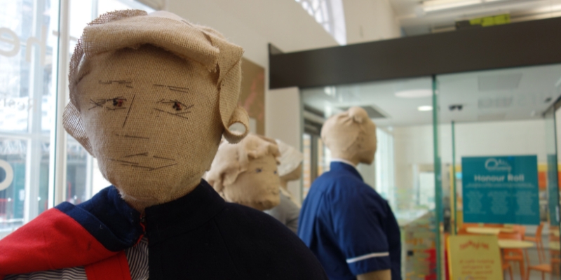 Up close image of the shoulders and head of a burlap mannequin dressed in a blue and red cape and a striped blue dress. In the background three other mannequins are partially visible in side view - one is more obvious and wearing a blue nurses dress with one hand in the pocket.