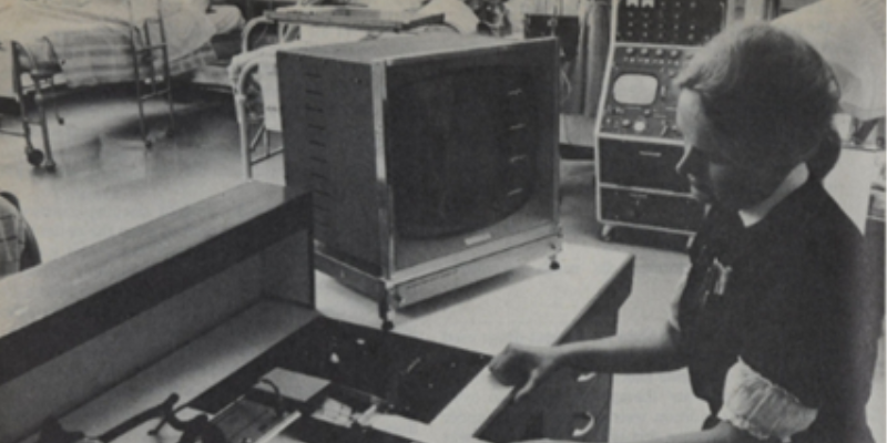 Black and white photo of a nurse in a hospital setting operating a piece of electrocardiogram equipment. A large monitor and control panels are visible.
