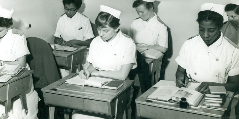 Black and white photograph of nurse students at desks, with piles of small books.