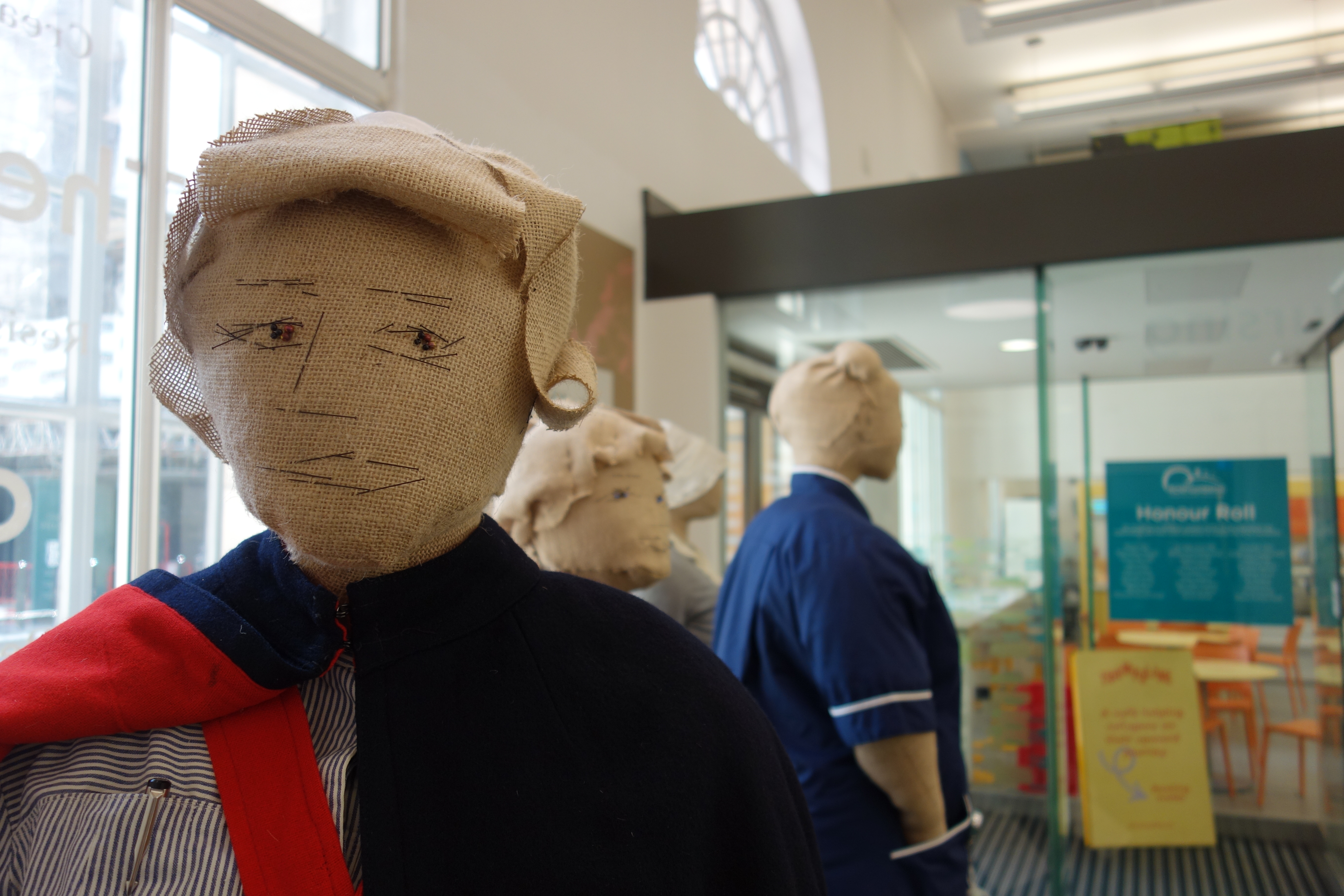 Up close image of the shoulders and head of a burlap mannequin dressed in a blue and red cape and a striped blue dress. In the background three other mannequins are partially visible in side view - one is more obvious and wearing a blue nurses dress with one hand in the pocket.