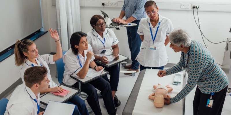 Nursing students around a table with a doll on it being taught by a lecturer