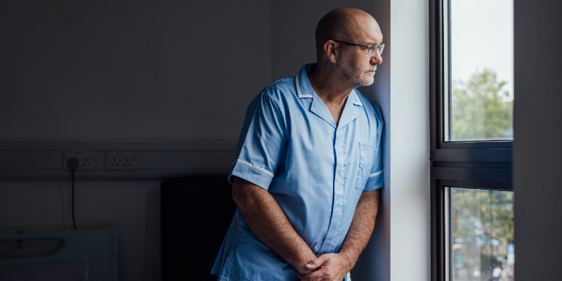 Older male nurse with a serious look on his face stands looking out of a large window 