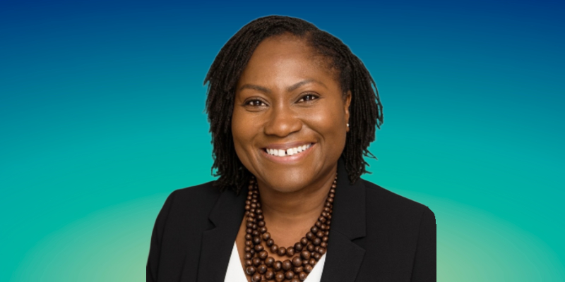 A woman smiles while looking directly into the camera. She is wearing a white top with a black jacket over the top and a necklace with lots of thick brown beads. There is a graded blue background behind her.
