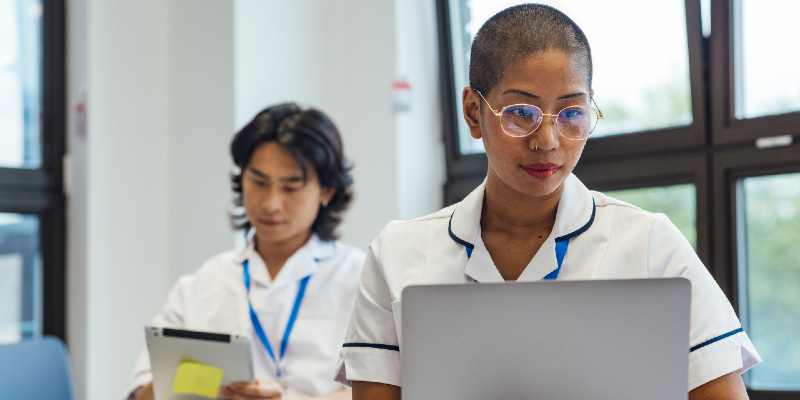 Two nursing students sat down, one using a laptop the other using a tablet