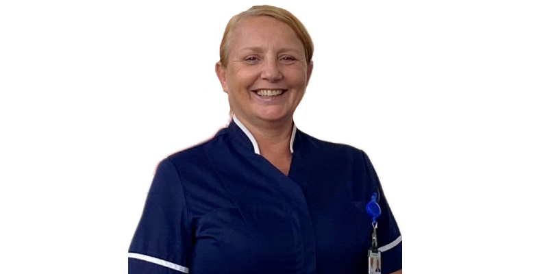 A woman is seen looking directly into the camera, smiling. She is wearing a navy nurse's uniform with white trim on the arms. A work pass is visible on her chest.