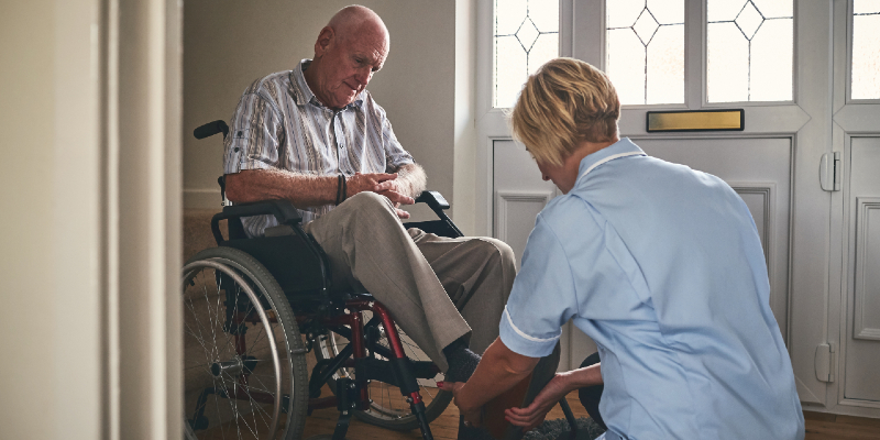 Female nursing staff member helping disabled senior man in wheelchair put his shoes on.