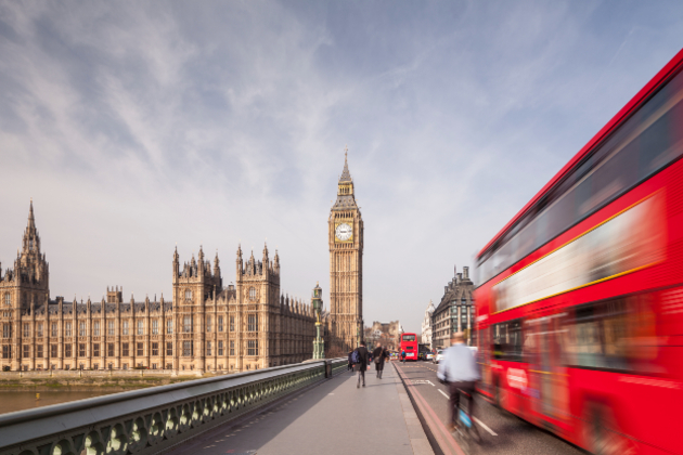 A view of the Palace of Westminster, the meeting place of the House of Commons and the House of Lords, the two houses of the Parliament of the United Kingdom. The picture has been taken from a bridge. Pedestrians walk ahead and a cyclist and blurred London bus pass on the right.