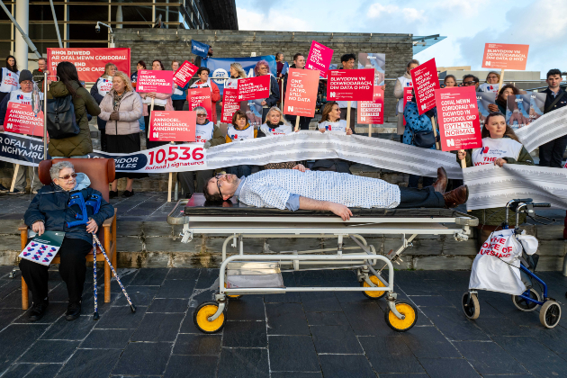 A group of people are shown protesting against corridor care outside the Senedd, the Welsh Parliament. In the centre in the foreground a man is shown lying down on a hospital trolley. He is surrounded by medical staff, members of the RCN and BMA, holding pink placards in English and Welsh