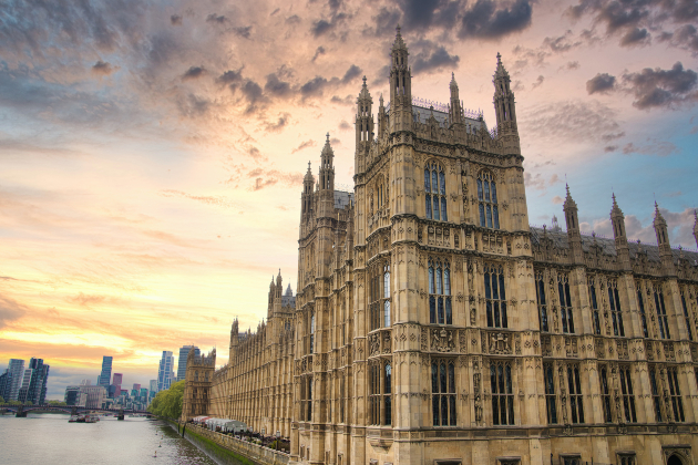 A view of the Houses of Parliament in Westminster in London. The River Thames can be seen towards the left of the photo, with other London skyscrapers visible. There is a dramatic sky behind with clouds and a sunset.