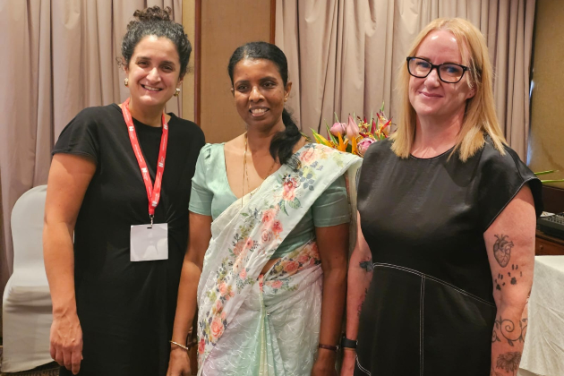 Two women smiling and dressed in black stand between a woman smiling in a pale green sari