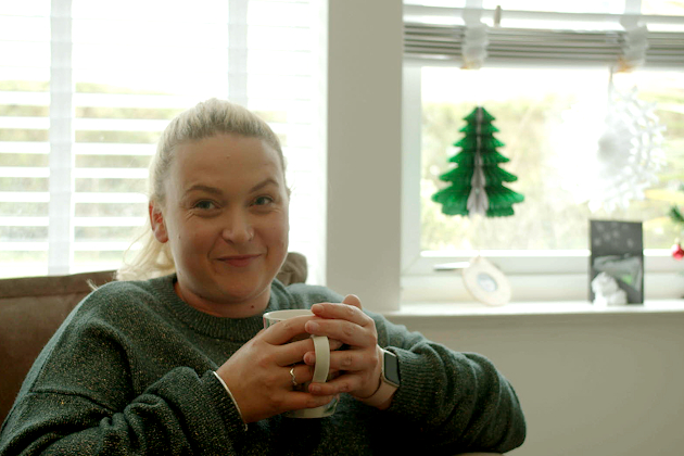 A woman smiles at the camera wearing a green jumper, holding a mug with a Christmas tree in the background