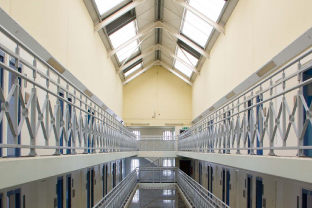Multiple floors of a prison are shown. Blue cell doors are seen among cream walls. Light floods in from skylights above and there is a metal barrier around the walkways and a mesh net is hung between the barriers on one of the floors.