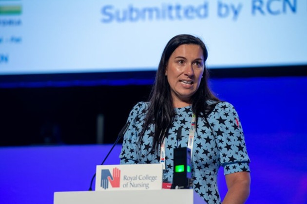 Francesca Steyne, a male fertility specialist nurse, is shown speaking at RCN Congress. She is wearing a light blue top with black stars patterned all over it. She stands at a lectern with a big screen behind her and microphones and the RCN logo in front of her. She has long, dark brown hair and her face is turned slightly towards the camera
