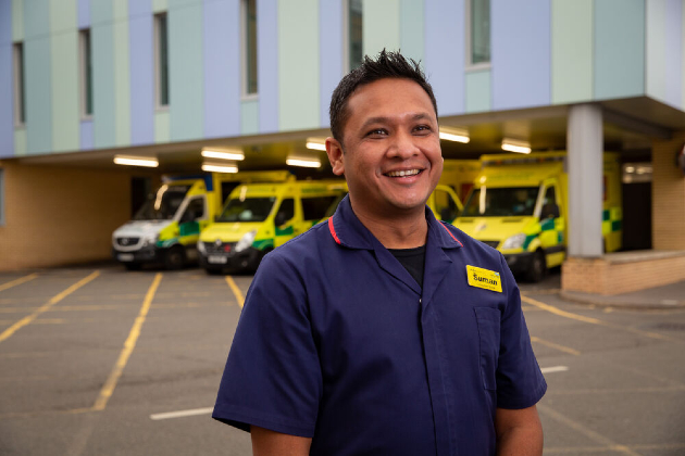 A nurse stands outside a hospital, seen from the waist up, with ambulances parked in a bay visible behind him. He is wearing a navy uniform with red trim and has a yellow name badge on. He is smiling, and looking off camera.