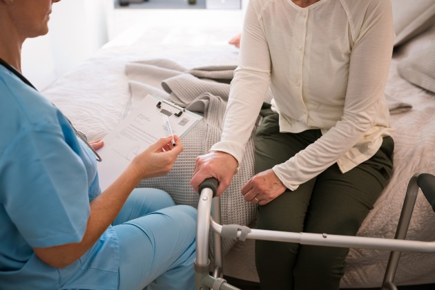 Nurse conducts a home equipment assessment with older woman seated on bed, with hand on a zimmerframe