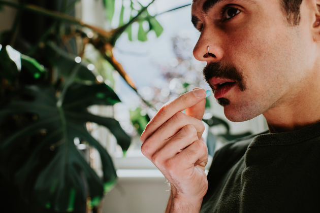 A man is seen holding a blue pill near his mouth, about to take the medicine. He looks around 30 years old, and has dark hair, a moustache and a nose ring. He is inside and in the background house plants can be seen, with light flooding into the room.