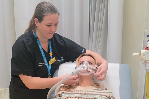 Karen in blue nurses uniform fitting a CPAP machine onto a patient who is lying on a bed