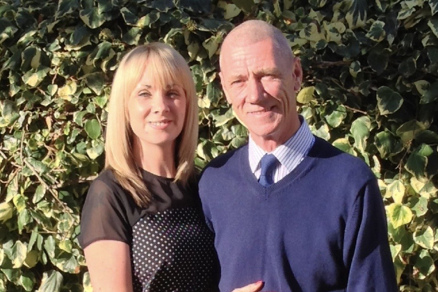 A woman and her dad are seen standing together. They are outside with sun on their faces and the leaves of a bush or hedge behind them. She has blonde, shoulder-length hair and wears a black top. He has a shaved head and wears a striped shirt with a blue tie and a jumper. They are both smiling.