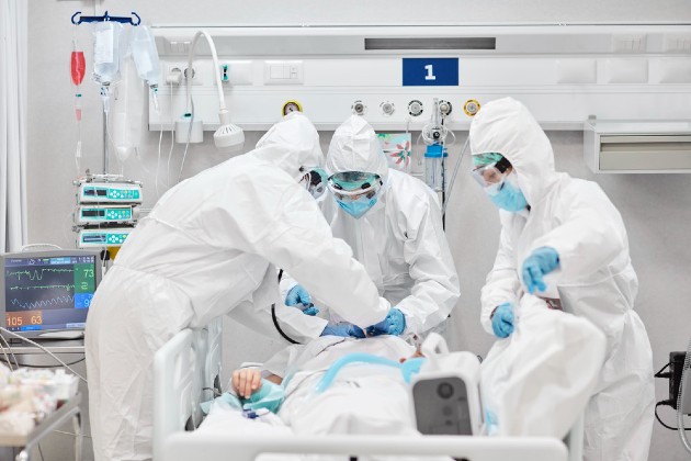 Three nurses in full PPE attend to a patient in a hospital bed in ICU