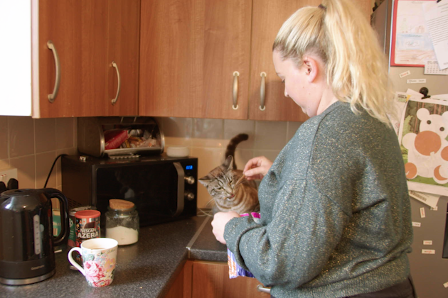 A woman is seen in profile, standing in a kitchen while making a hot drink. She is wearing a green jumper and petting a cat, which stands on the worktop.